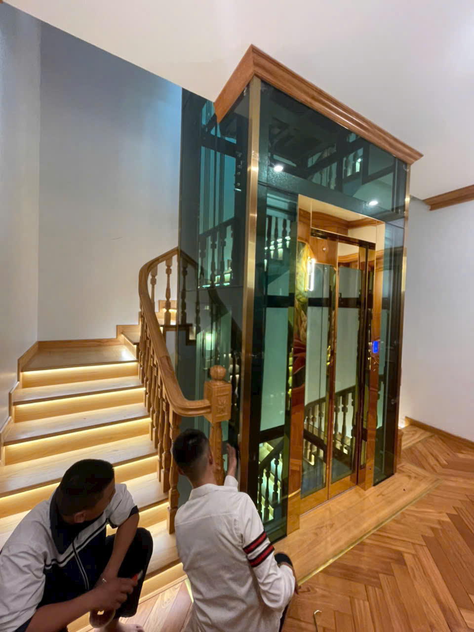 Thang máy gia đình kính khung gỗ đang được kỹ thuật viên lắp đặt cạnh cầu thang gỗ có đèn LED